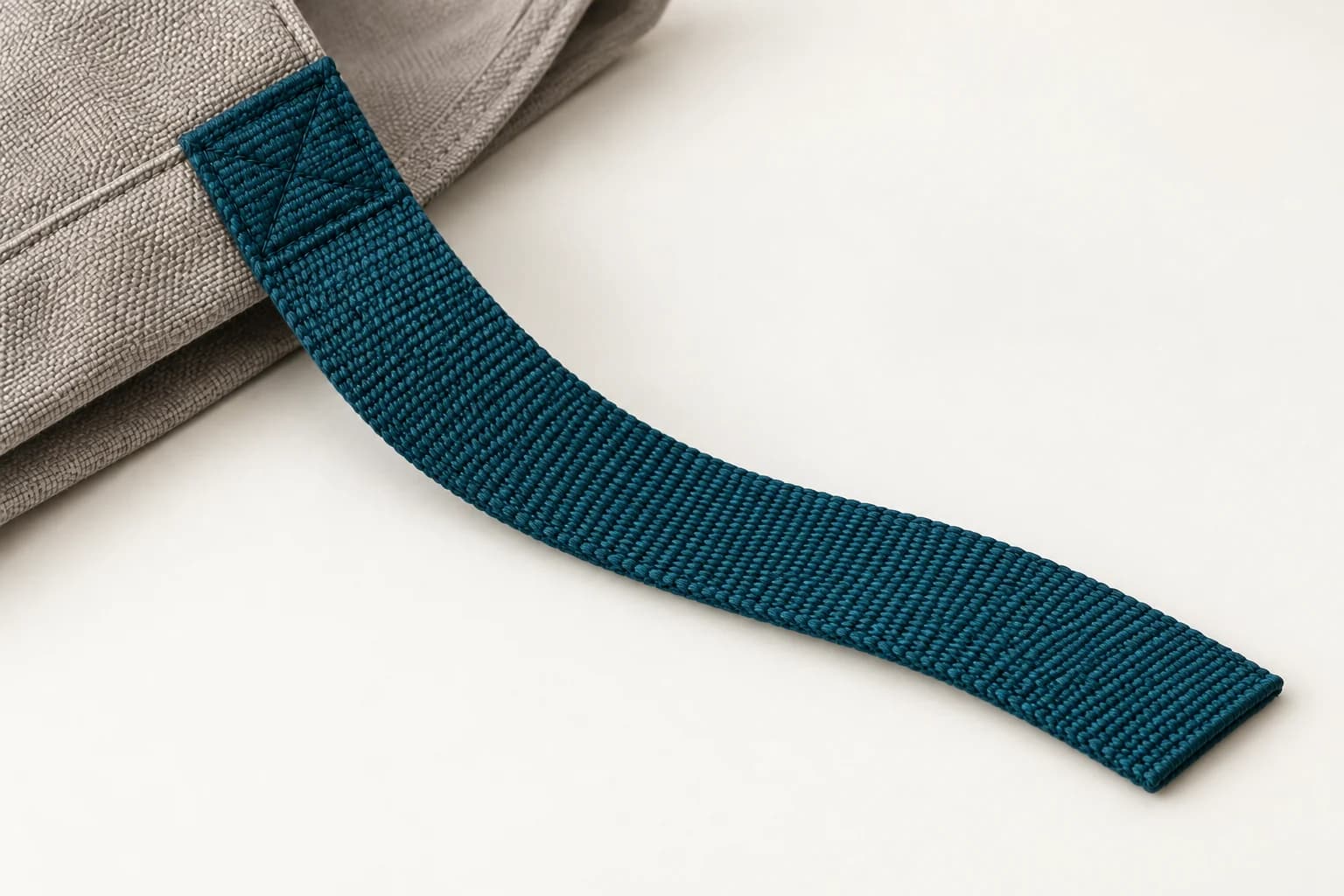 A teal flat woven strap stitched onto grey bag fabric with its free end visible